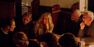 Mennesker samlet i et lokale oplyst af levende lys. De diskuterer og samtaler indgående.