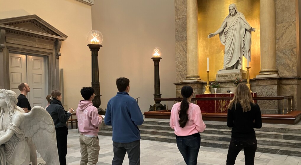 En præst uden præstekjole står sammen med fem studerende og betragter Thorvaldsens jesusskulptur i Københavns Domkirke. De studerende har pen og blok i hænderne og tager noter eller skitser.