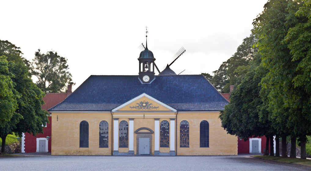 Kastelkirken er en gulkalket barokkirke med et klokketårn midtfor på taget omgivet af grønne trækroner og bag kirkens tag anes Kastellets møllevinger
