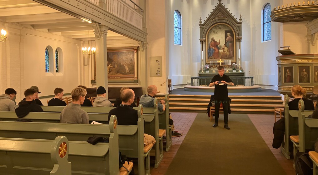 I en københavnsk kirke står en præst i en t-shirt og en bog i hænderne. Han er i færd med at forklare noget til de studerende, der lytter med fra kirkebænkene i forgrunden