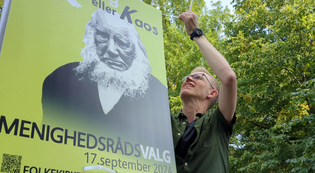 En sognepræst i civilt tøj er klatret op på en stige, hvorfra han hænger en valgplakat op.