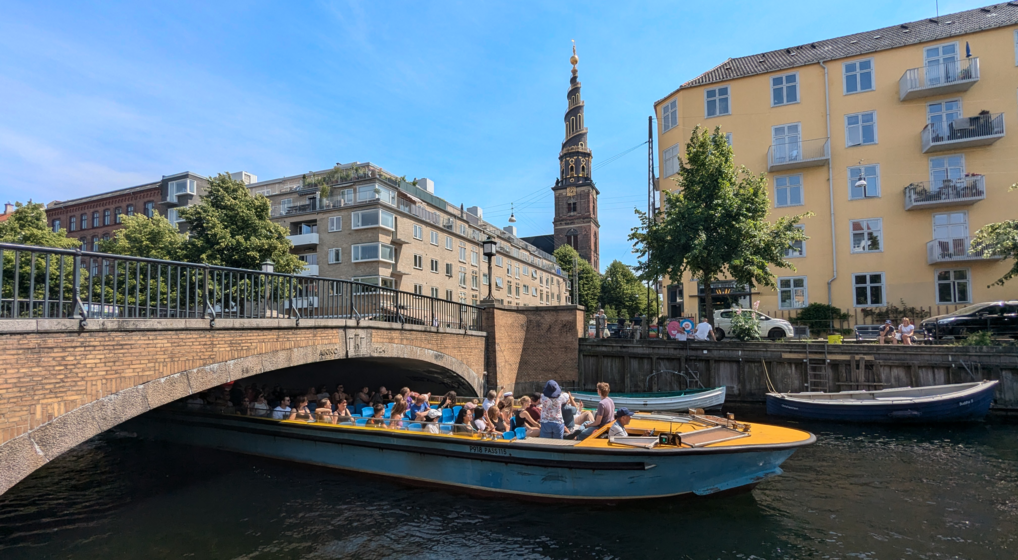 En turistbåd sejler under en bro i Københavns Kanal med Vor Frelsers Kirkes tårn i baggrunden midt i billedet