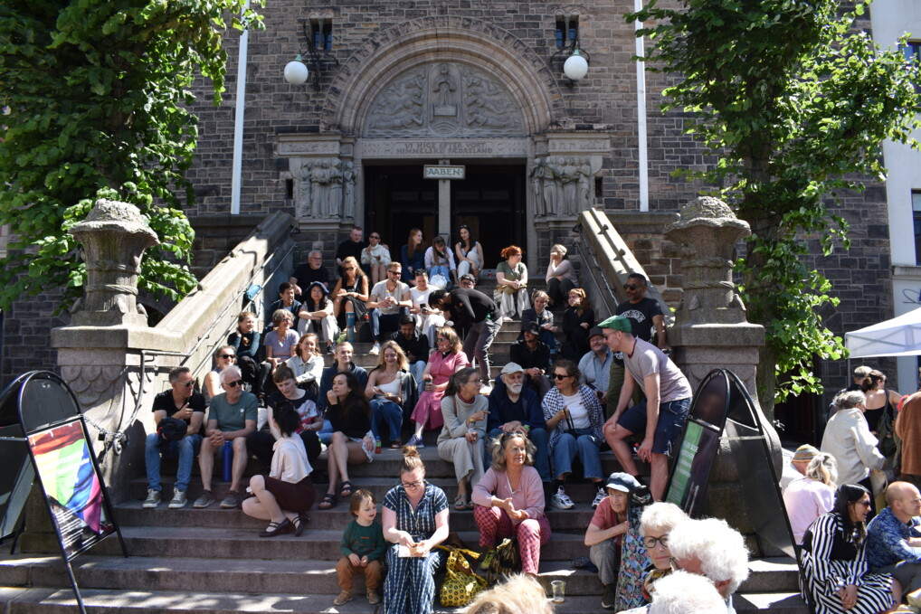 En masse mennesker sidder på trappen foran Eliaskirken på Vesterbro i København. Solen skinner og folk smiler