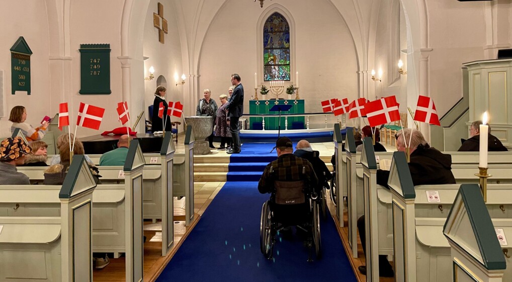I et kirkerum pyntet med danske flag sidder mennesker på kirkebænkene eller i kørestol i gangen