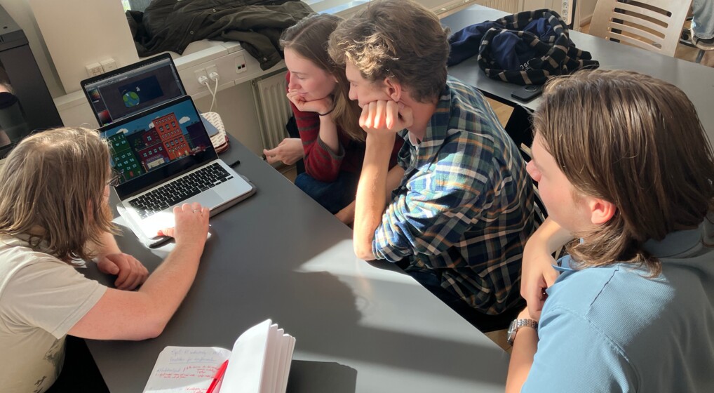 Tre studerende sidder omkring en computer med deres underviser og ser på et farverigt computerspil de selv har udviklet i skoletiden