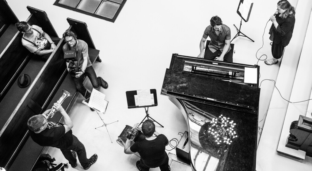 Et sort-hvid billede med et flygel placeret lidt til højre for midten med jazzmusikere omkring. Koncerten foregår på kirkegulvet i Eliaskirken på Vesterbro, København.