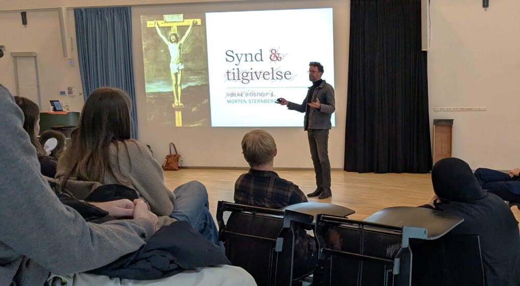 En præst i civil taler til en gymnasieklasse foran et lærred med overskriften "Synd og tilgivelse"