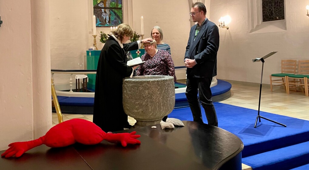 Tre personer og en præst står samlet om døbefonten, hvor en kvinde med kort, gråt hår er i færd med at blive døbt