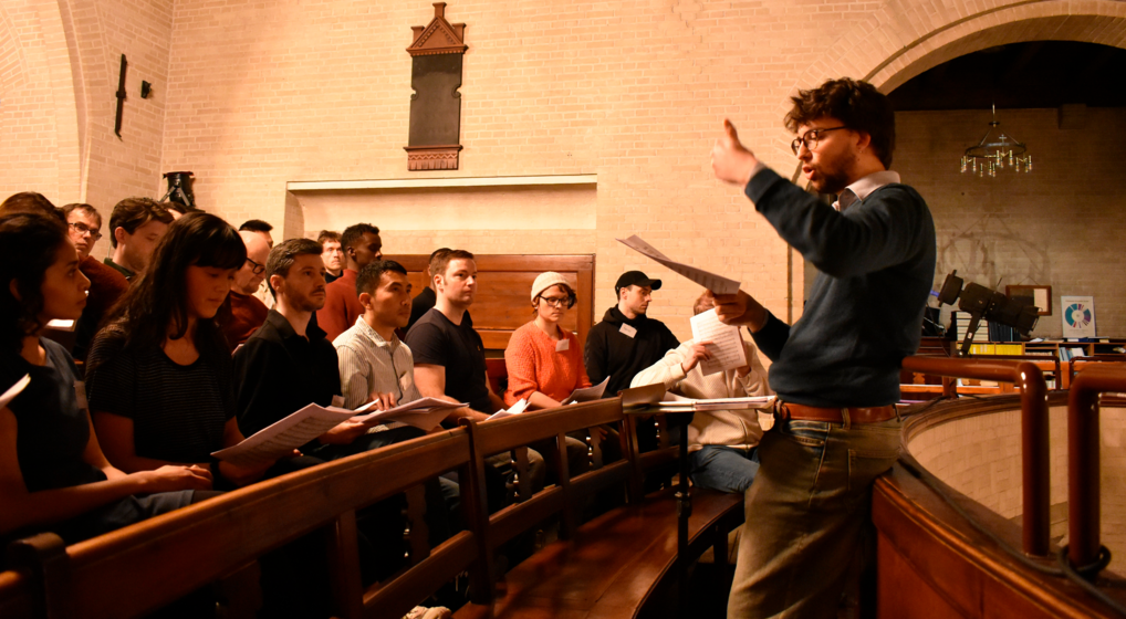 A choir rehearsing in a church. The conductor waves his hands as he gives out instructions.