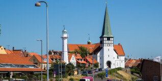 En hvidkalket kirke med et højt, spidst, grønt-irret tårn ligger på toppen af en forhøjning med udsigt over Rønne Havn. Himlen er blå på en sommerdag og vinden tager i de blafrende flag langs vejen for foden af kirken