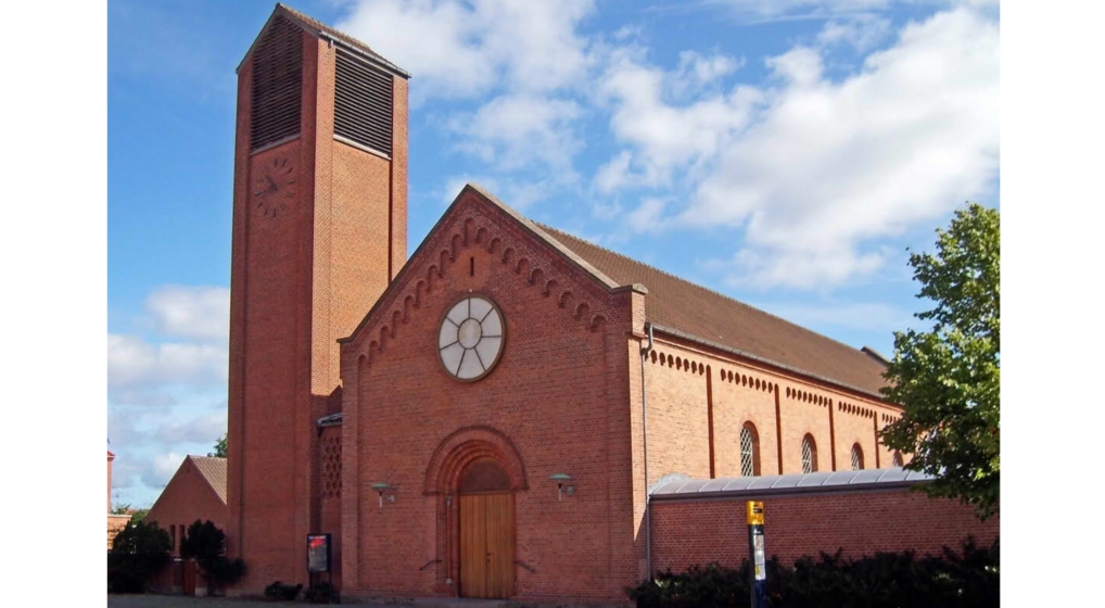 Kastrup Kirke er en murstenskirke fra slutningen af 1800-tallet. Tårnet står ved siden af kirkeskibet og har en urskive. Billedet er taget en sommerdag med små hvide skyer på den blå himmel.